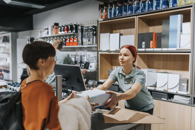 Female customer giving appliance box to sales clerk at checkout counter in electronics store