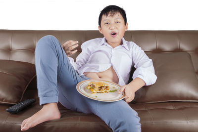 Portrait of boy eating burger while sitting on sofa against white background