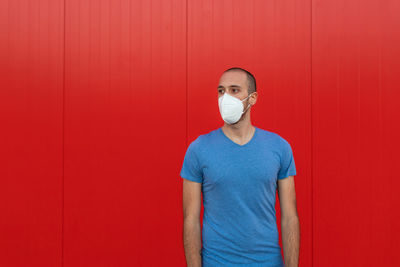 Young man wearing respiratory mask while looking away near colorful red wall during coronavirus pandemic