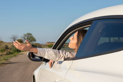 Woman with eyes closed leaning out of car window with arm outside enjoying fresh breeze of nature