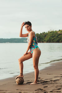 Full body side view of young female in colorful swimsuit and sunglasses stepping on dried coconut on sandy beach near sea in summertime in costa rica