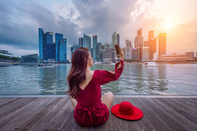 Rear view of woman taking selfie by lake against city