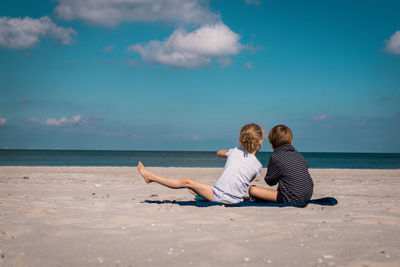 Rear view of siblings sitting at beach against blue sky