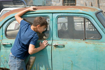 A boy 11 years old looks out the window of a rusty car in a dump of abandoned cars on a sunny day.