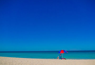 Single male relaxes on an empty sandy beach under a colorful parasol and clear blue sky
