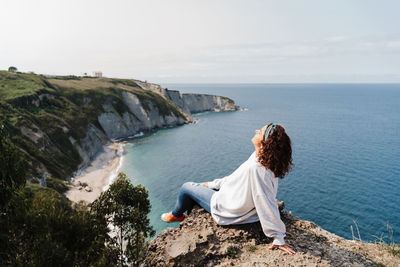 Woman sitting on cliff by sea against sky