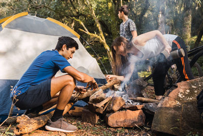 Multiethnic young explorers kindling smoking bonfire near tent during hike on bicycles in woods