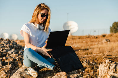 Young woman wearing sunglasses with laptop sitting on land against sky