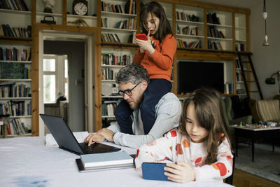 Daughter using smart phone by father working on laptop with girl sitting on shoulders at home