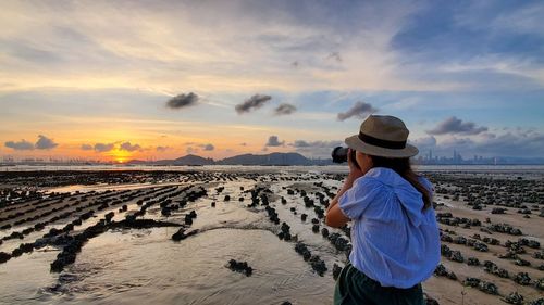 Rear view of woman photographing at beach against sky during sunset