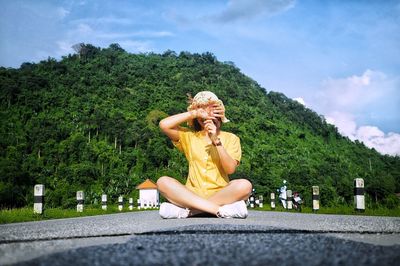 Full length of woman covering and holding stems over face while sitting on road against sky