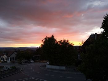 Road by trees and buildings against sky during sunset