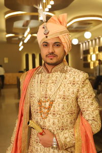 Portrait of young groom in indian royal wedding attire standing against illuminated ceiling