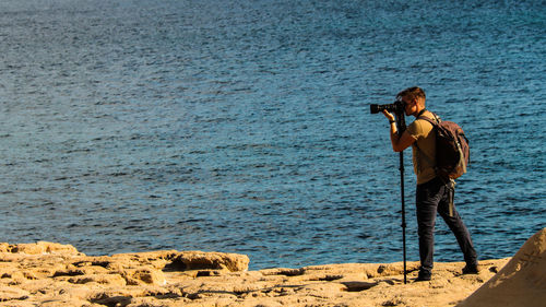 Full length of man photographing through camera on rocks by sea