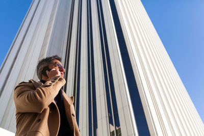 Young male in stylish outfit and sunglasses talking on mobile phone while standing on city street