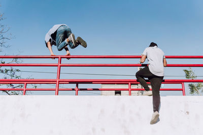 Low angle back view of fearless unrecognizable male friends jumping above metal railing in city while performing parkour stunt on sunny day