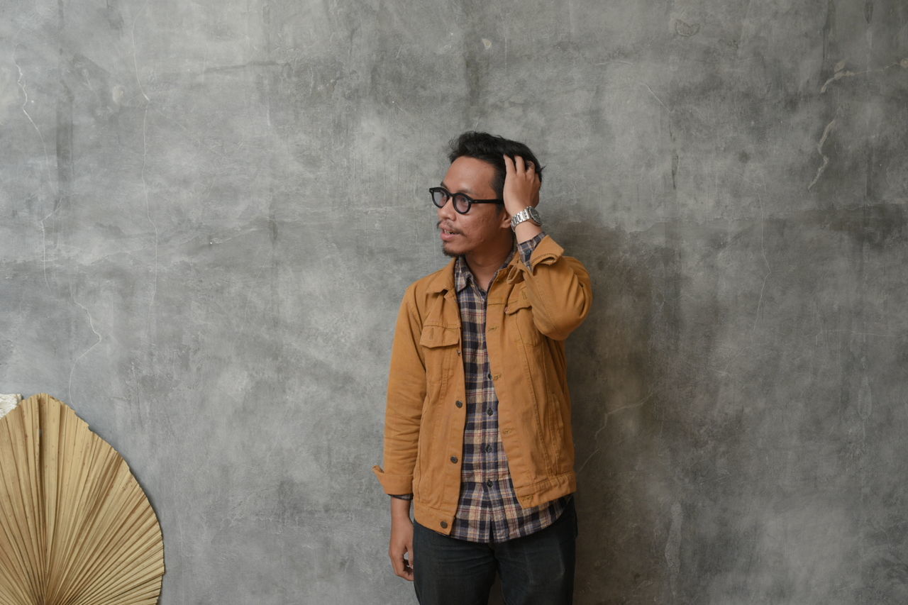 one person, adult, glasses, standing, wall - building feature, men, casual clothing, spring, copy space, eyeglasses, young adult, looking, front view, person, clothing, fashion, architecture, lifestyles, photo shoot, gray, cool attitude, hands in pockets, portrait, three quarter length, yellow, emotion, music, looking away, city, smiling, indoors