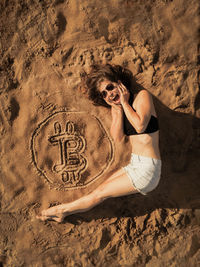 Portrait of young woman lying down by bitcoin symbol on sand at beach