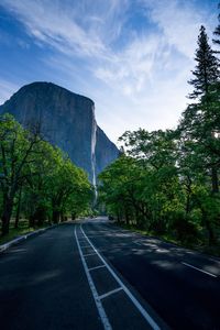 Road amidst trees leading towards rocky mountain against sky