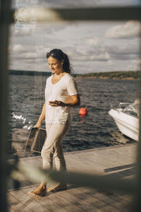 Full length of woman with mobile phone and laptop walking on patio by lake