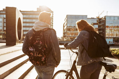 Teenage boy walking with friend holding bicycle in city during sunny day