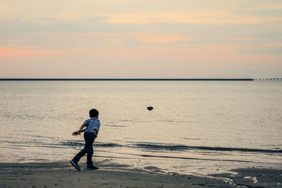 Rear view of boy throwing stone into sea during sunset