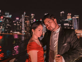 Portrait of man standing with girlfriend against river in illuminated city at night