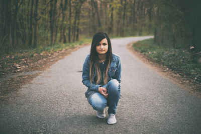 Portrait of young woman crouching on road with trees in background