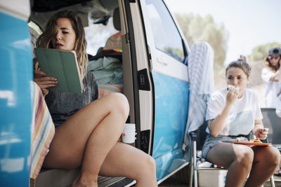 Woman reading tablet pc while female friend eating by camper van