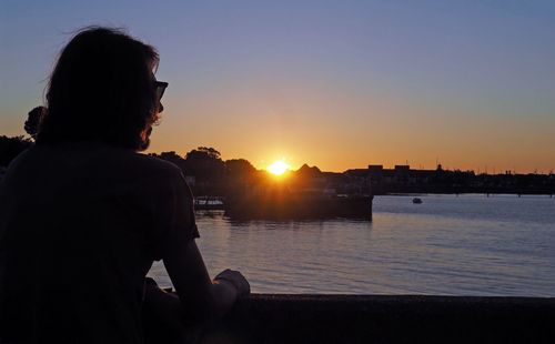 Rear view of woman standing by river against sky during sunset