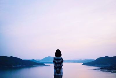 Rear view of woman standing by river and mountains against sky