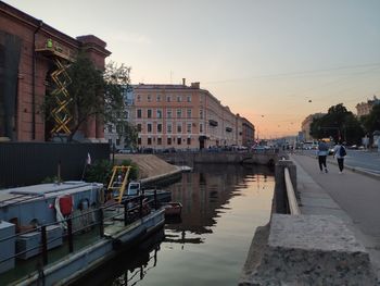 People in canal by buildings against sky during sunset