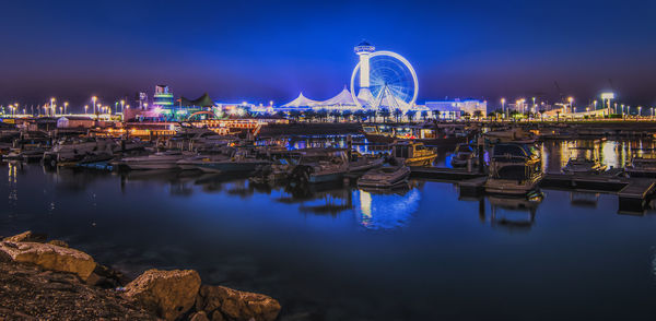 Illuminated amusement park by water front against sky at night