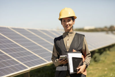 Portrait of smiling female maintenance engineer holding smart phone and documents while standing near solar panels in fi