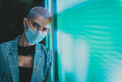 Portrait of young woman with shaved head wearing mask standing against abstract backgrounds