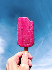 Close-up of hand holding pink popsicle with a bite missing against a sky background 