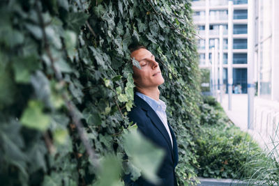 Businessman with eyes closed leaning on plant wall