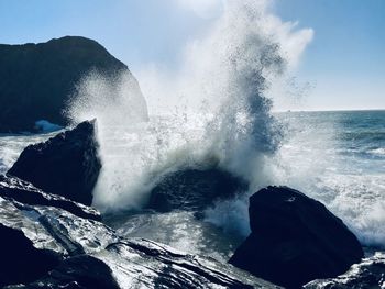 Waves splashing on rocks at shore against sky