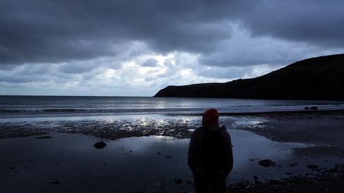 Rear view of person standing at beach against cloudy sky