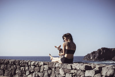 Healthy fit woman with tattoos sits by ocean with in sun with tiny dog