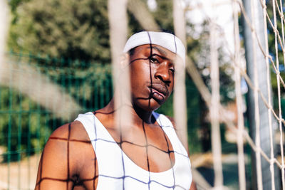 Serious african american male athlete in white activewear standing near net on playground in summer and looking at camera