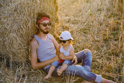 Father and son in t-shirts sitting next to a haystack on a sloping field during sunset