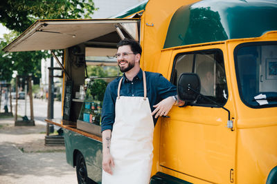 Happy young owner looking away while standing against commercial land vehicle on street