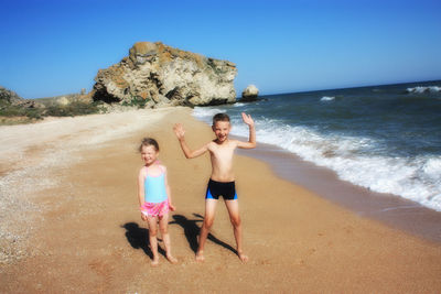 Full length of siblings standing on shore at beach against clear sky