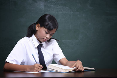 Schoolgirl studying at table against blackboard