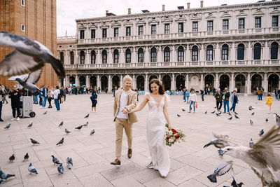 Full length of bridegroom holding hands walking against building