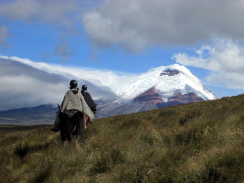 Rear view of people riding horses on grassy field against cotopaxi