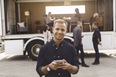 Portrait of smiling businessman holding mobile phone with colleagues and portable office truck on road in background