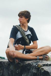 Young man looking away while sitting on rock against sky