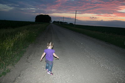 Rear view full length of girl standing on road amidst grassy field during sunset